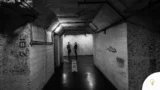 Moody black and white photo of two people in a dimly lit underground corridor